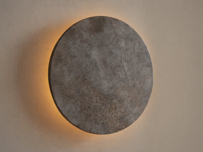 Odette Outdoor Sconce: Oval dark grey natural stone sconce on a beige wall, emitting warm golden LED light from behind.