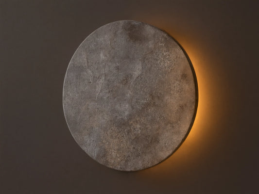 Odette Outdoor Sconce: Oval dark grey natural stone sconce on a beige wall, emitting warm golden LED light from behind.