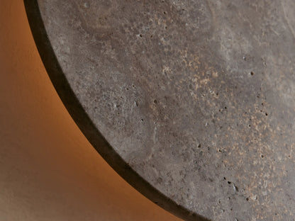 Odette Outdoor Sconce. Circular natural stone sconce, mottled grey with brown and lighter gold tones, on a beige wall.