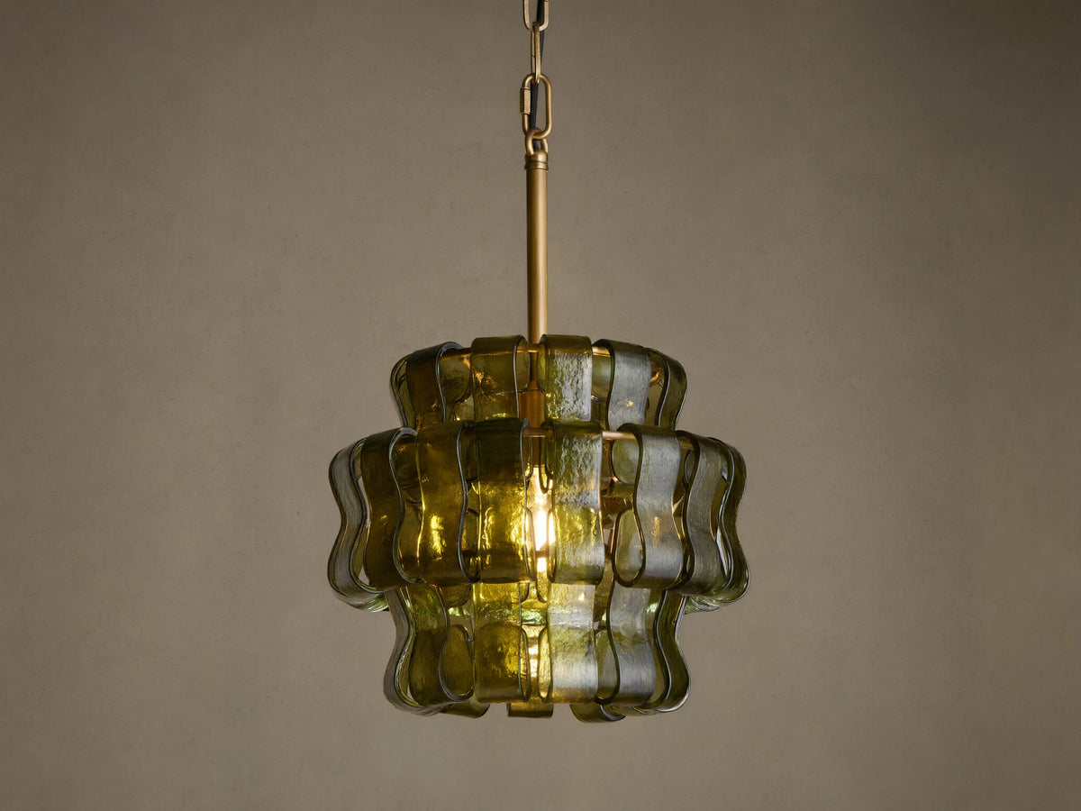 Ghiaccio Pendant, a tiered dark green wavy curved glass pendant light with brass chain, glowing warmly against a neutral wall.