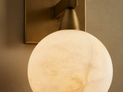 Front view of a brass wall sconce with a square backplate and round milky white glass shade, on a light wall next to a mirror.