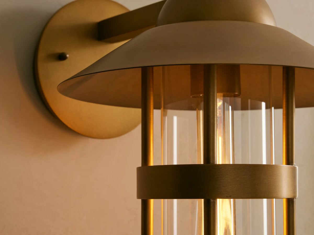 Hyannis Outdoor Sconce, a nautical-style brass lantern with clear glass, mounted on a light wall, left of a mirror.