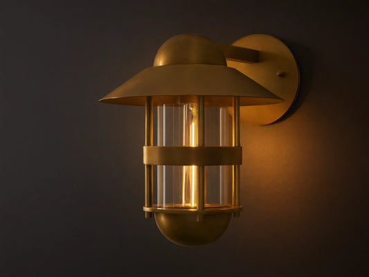 Illuminated Hyannis Outdoor Sconce, antique brass, nautical style with glass housing and visible bulb, on a light wall.