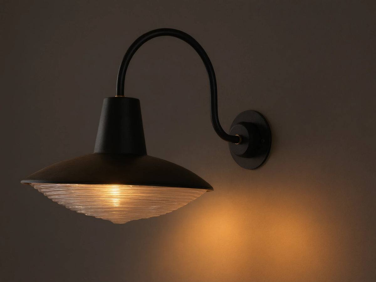 Close-up of Bennett Outdoor Sconce, black wall-mounted with a curved arm. Ribbed clear glass diffuser glows with warm light.