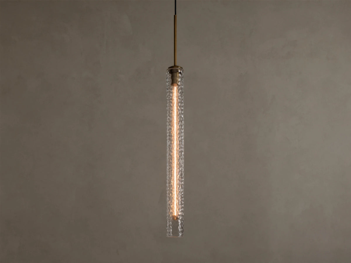 Vertical view of a lit pendant light with a clear textured glass cylinder, a warm filament bulb, and a brass-colored top fixture.