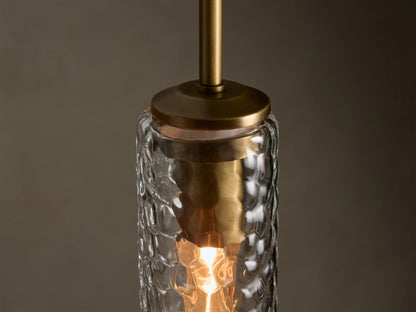 Clear glass pendant light, featuring a vertical hammered cylindrical shade, a brass top, and a warm glow.
