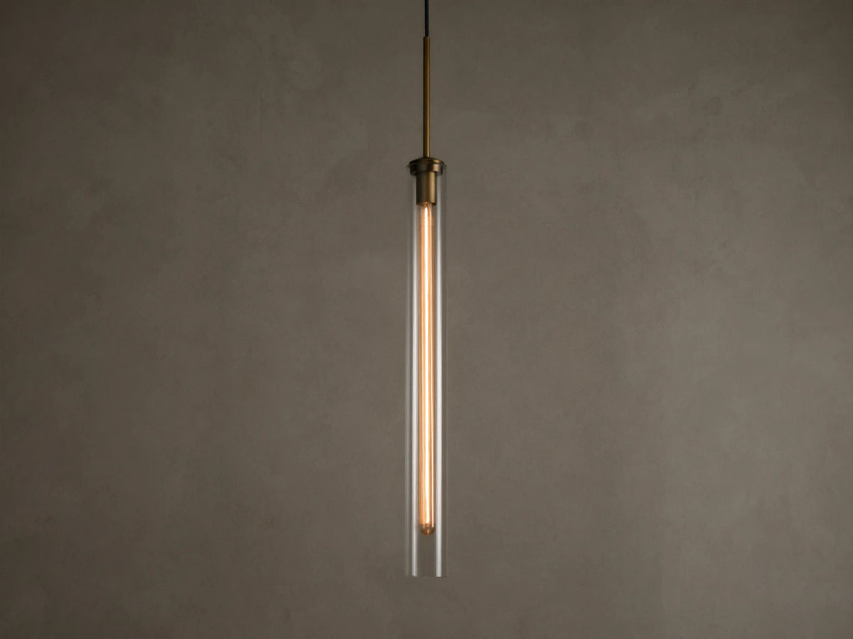 Front view of a modern brass and clear glass pendant light fixture with a long, illuminated filament bulb inside.