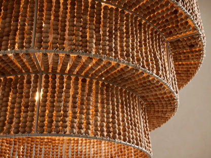 Kash Chandelier, a tiered light brown wooden bead chandelier with an ivory iron frame, hanging vertically against a beige wall.