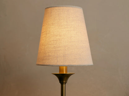 Saige Sconce, close-up of its illuminated off-white linen conical shade and dark brass finish metal body against a neutral wall.