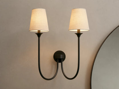 Saige Sconce with two off-white shades & dark metal arms, illuminated on a warm-toned wall next to a mirror.