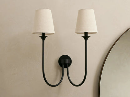 Saige Sconce: Two black metal wall sconces with off-white conical fabric shades on a beige wall, beside a round mirror.