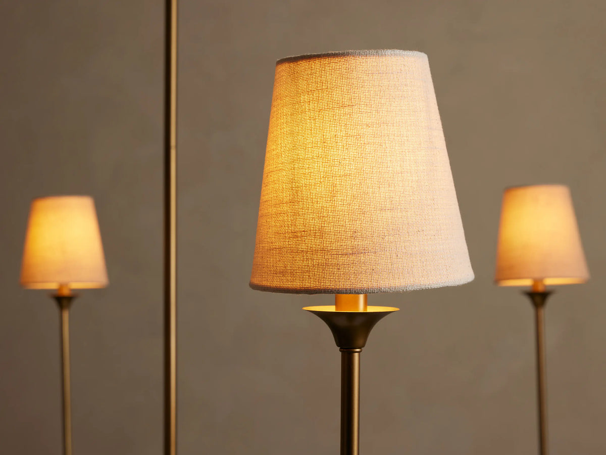 Close-up of Saige Round Chandelier's warm brass-finished light with an illuminated off-white fabric conical shade. Two blurred lights in background.
