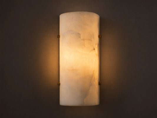 Wall-mounted cylindrical sconce made of alabaster, softly glowing with warm, inviting light against a beige textured wall.