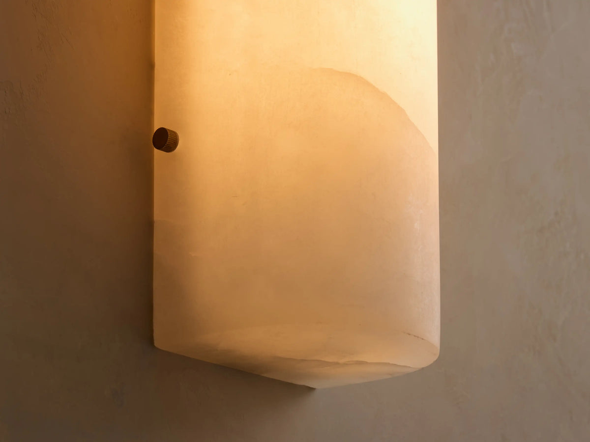 A cylindrical white alabaster wall sconce mounted vertically on a light beige textured wall, adjacent to a mirror.