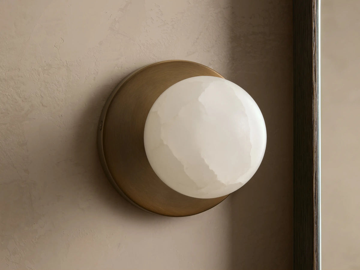 A round, white alabaster wall sconce with a soft glow, set against a textured beige wall. The base is a matte brass finish.