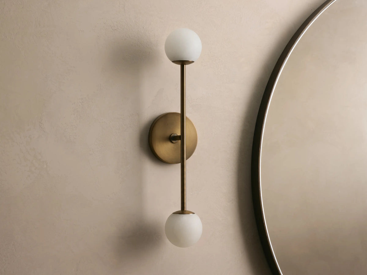 Bronze wall sconce with two frosted glass globes, vertically aligned. Mounted on a light beige wall, near a round mirror.
