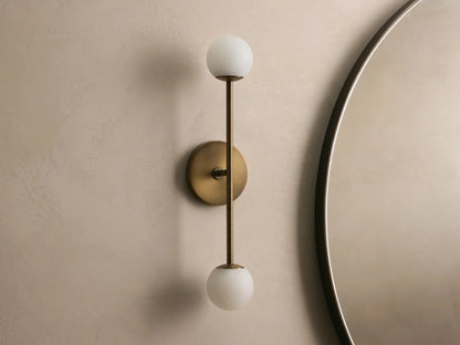 Bronze wall sconce with two frosted glass globes, vertically aligned. Mounted on a light beige wall, near a round mirror.