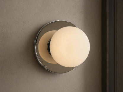 A round wall sconce with a frosted glass globe emits warm light, mounted on a smooth circular metal base, against a beige wall.
