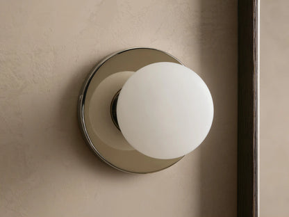 A round wall sconce with a frosted glass globe emits warm light, mounted on a smooth circular metal base, against a beige wall.