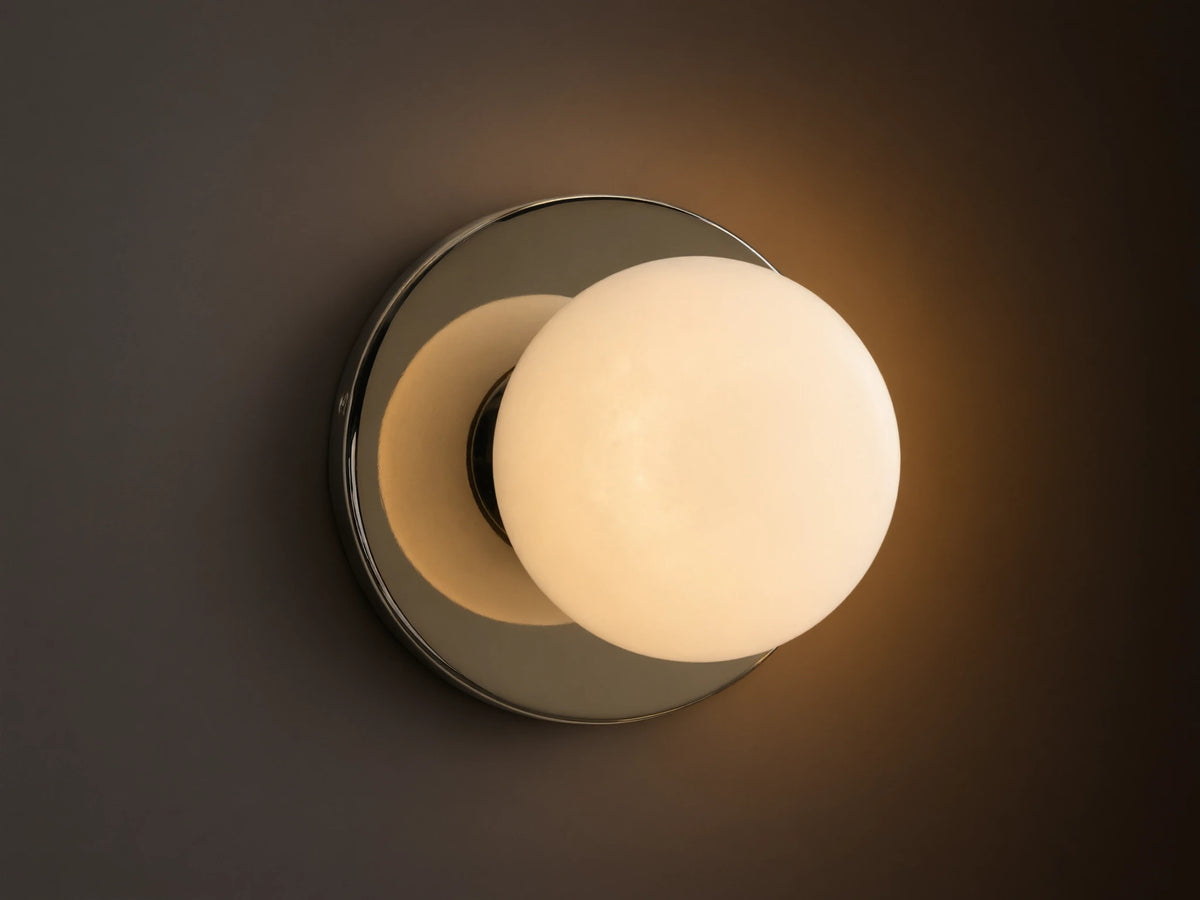 A round wall sconce with a frosted glass globe emits warm light, mounted on a smooth circular metal base, against a beige wall.
