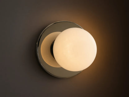 A round wall sconce with a frosted glass globe emits warm light, mounted on a smooth circular metal base, against a beige wall.