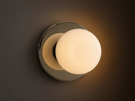 A round wall sconce with a frosted glass globe emits warm light, mounted on a smooth circular metal base, against a beige wall.