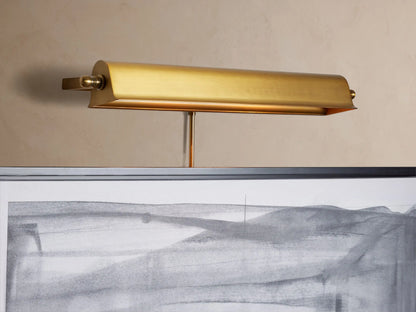 Angled close-up of an antiqued brass Tremont Picture Light Sconce, glowing warmly, mounted on a light beige wall.