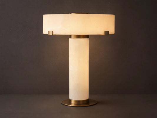 Chayla Table Lamp with glowing white alabaster shade, cylindrical alabaster base, and antique brass accents on a wooden table.
