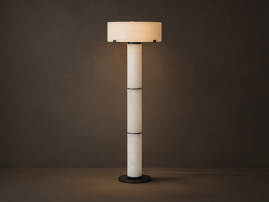 Chayla Floor Lamp with a cylindrical alabaster base, bronze accents, and a round, white lampshade, standing on a wooden floor.
