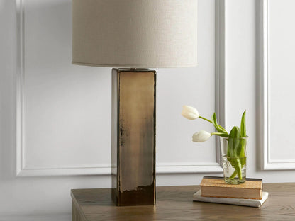 Adrano Table Lamp with antique copper finish base and natural linen drum shade on wooden surface, near books and tulips.