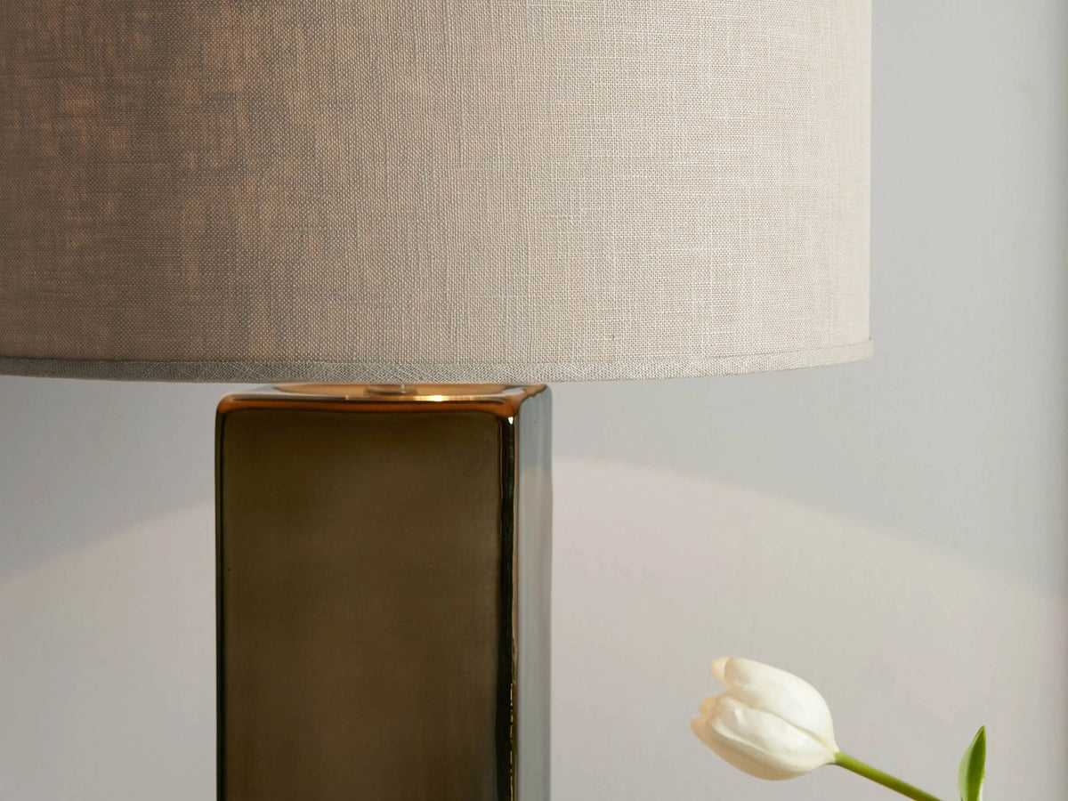 Adrano Table Lamp on a wooden surface, featuring a gold-toned rectangular glass base and a natural linen drum shade. Books and a vase with tulips are beside it.