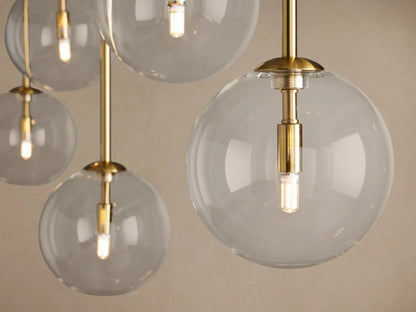 Roscoe Linear Chandelier: Close-up of clear glass spheres, brass-plated rods, and glowing LED bulbs against a light wall.