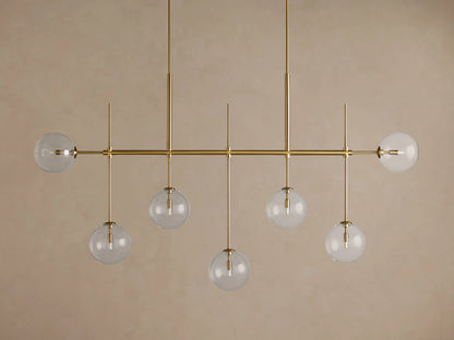 Roscoe Linear Chandelier: 7 illuminated clear glass globe lights on a brass-finished horizontal bar. Angled view.