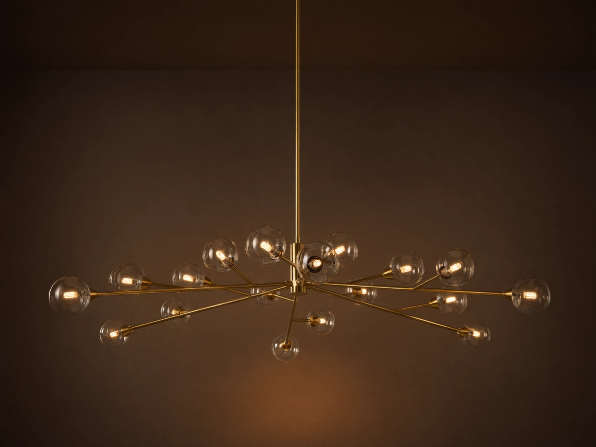 Apollo Round Chandelier: Brass-toned arms with many illuminated clear glass globe bulbs, suspended against a textured wall.