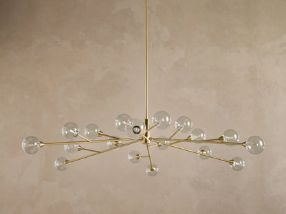 Apollo Round Chandelier: Brass-toned arms with many illuminated clear glass globe bulbs, suspended against a textured wall.