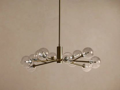 Apollo Round Chandelier, brass frame, clear spherical bulbs glowing brightly, hanging against a warm brown wall.