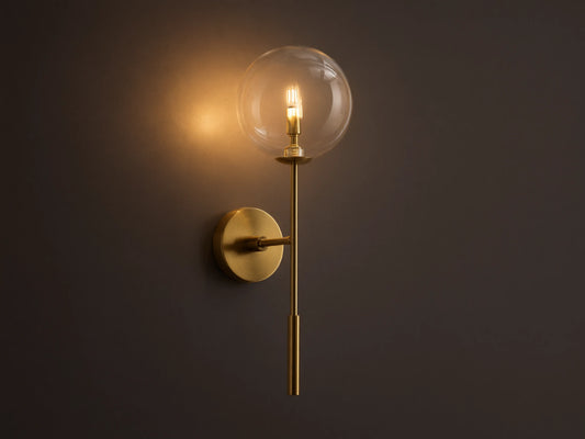 Front view of the brass Roscoe Sconce, featuring a clear glass globe with an illuminated LED bulb, mounted on a wall.