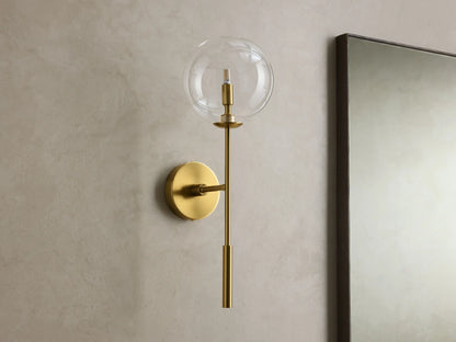 Front view of the brass Roscoe Sconce, featuring a clear glass globe with an illuminated LED bulb, mounted on a wall.