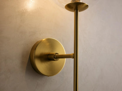 Front view of the brass Roscoe Sconce mounted on a wall, with a clear glass globe and linear rod.