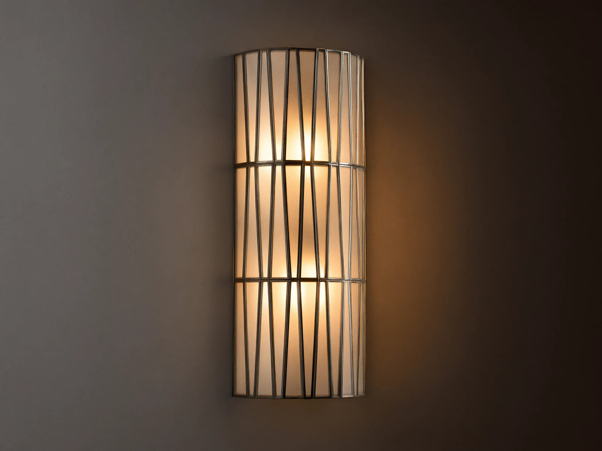 Jeunet Sconce with antiqued brass finish, features two glowing lights behind a cylindrical iron frame with vertical and diagonal lines.