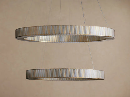 Jeunet 2-Tier Round Chandelier: Two illuminated, antiqued brass rings with vertical glass slats hang horizontally, glowing warmly.