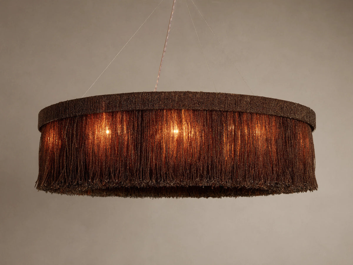 Romilly Round Chandelier, dark bronze beaded fringe, emitting a warm orange glow from internal lights, hanging against light wall.