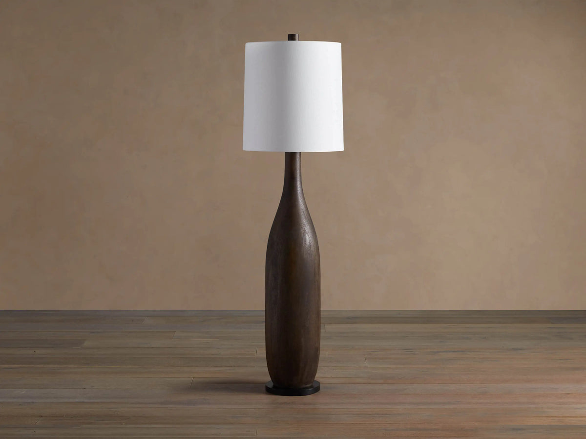 A tall Briar Floor Lamp with a dark espresso, slender wooden base and a glowing white linen cylinder shade on a wooden floor.