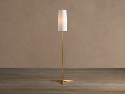 Lancia Floor Lamp with slender brass pole, triangular base, and tall, glowing white linen shade on a wood floor.