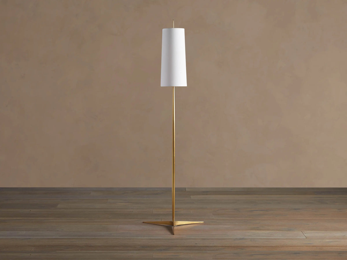 Lancia Floor Lamp with slender brass pole, triangular base, and tall, glowing white linen shade on a wood floor.