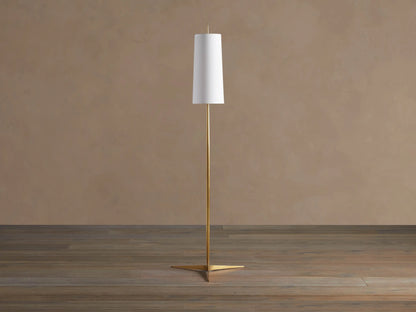 Lancia Floor Lamp with slender brass pole, triangular base, and tall, glowing white linen shade on a wood floor.