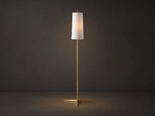 Lancia Floor Lamp with slender brass pole, triangular base, and tall, glowing white linen shade on a wood floor.