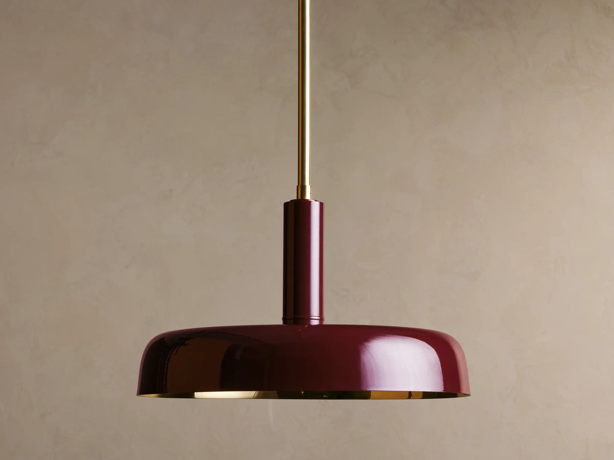 A maroon pendant light with a glossy finish, suspended from a brass rod, against a beige background. The light is off.