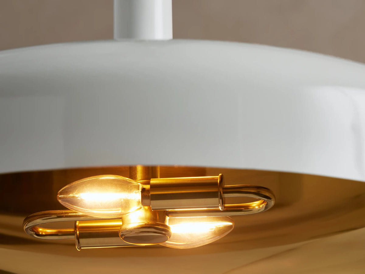 Bonham Pendant: white dome shade with a brass interior, suspended by a brass rod, against a light brown wall.