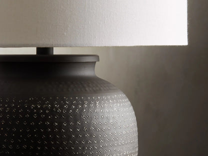 Howell Table Lamp with an embossed, dark powder-coated base and a wide, white cotton linen shade, on a wooden surface.